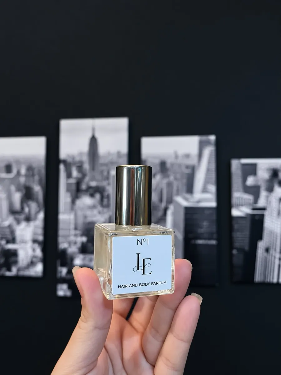Frasco LE E'ssence Nº1 Hair and Body Parfum, com tampa dourada, segurado na mão diante de fotografias de Nova York em preto e branco